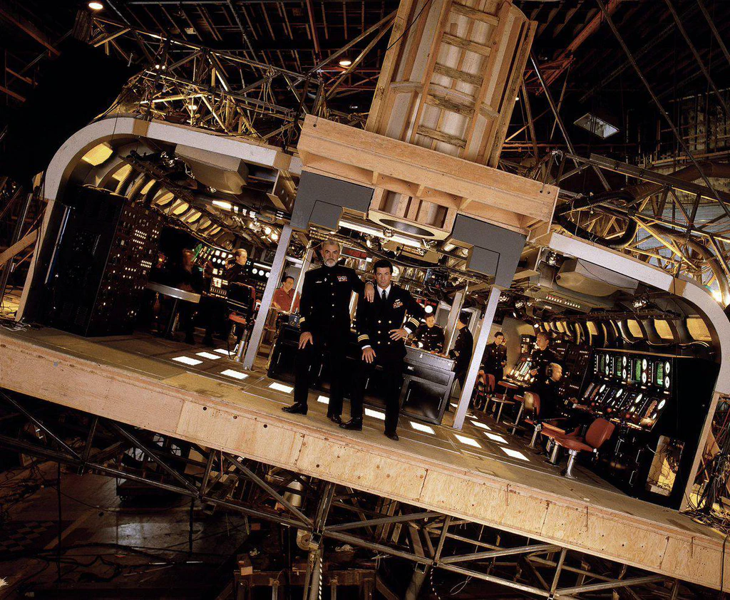 A behind-the-scenes view of a movie set designed to resemble a spaceship cockpit, with actors in military-style uniforms and multiple technical crew members working around it.