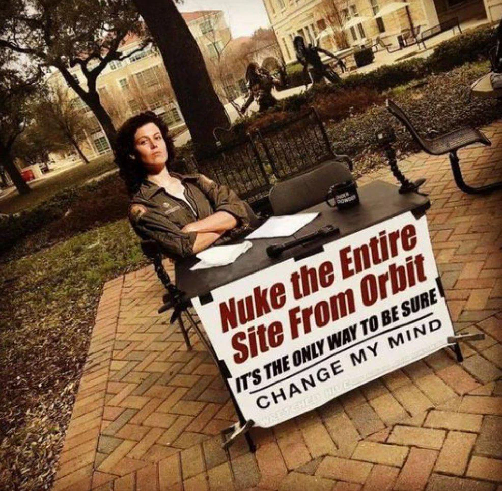 A woman sitting at a table outdoors with a sign that reads, "Nuke the Entire Site From Orbit. It's the only way to be sure. Change my mind."