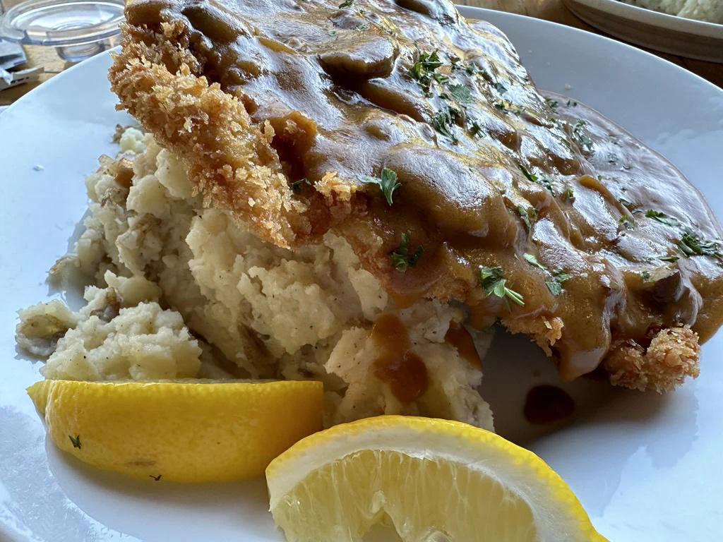 A plate with a breaded fried pork chop topped with gravy, served with mashed potatoes and two lemon wedges.