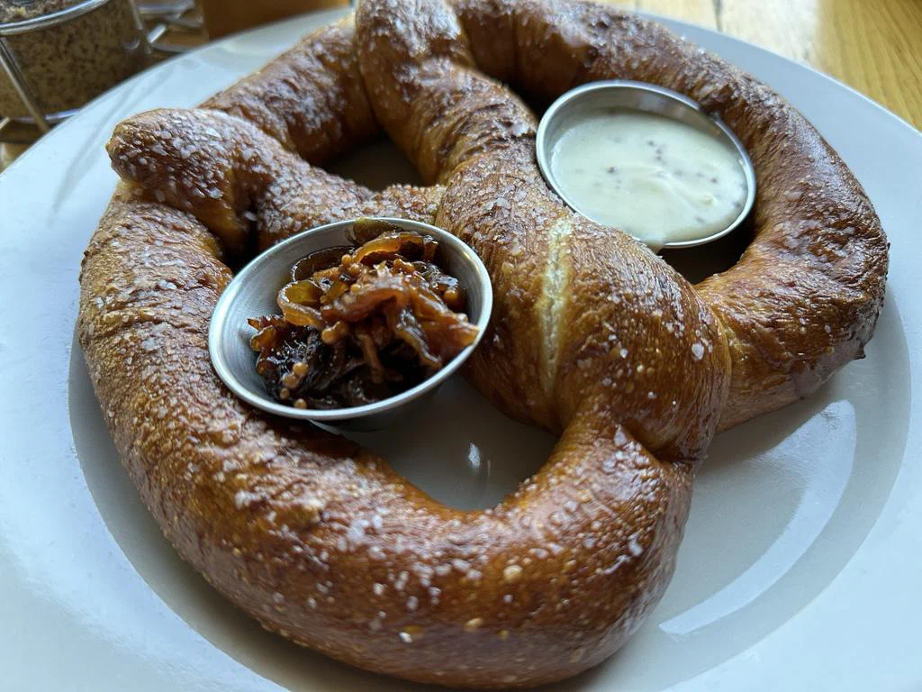 A plate of two large, crispy, golden-brown pretzels served with dipping sauces, one appears to be a creamy white sauce and the other a caramelized onion or sweet relish.